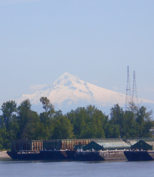 Large, pointed snow peak rises above four barges on the Willamette River