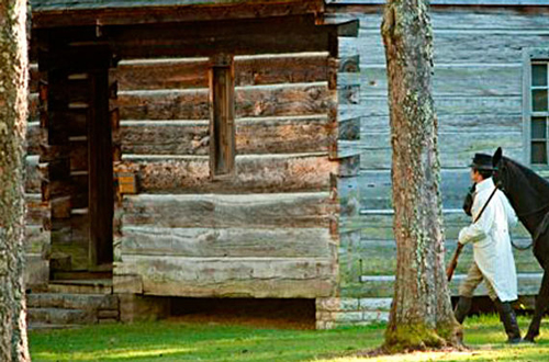 A solitary man leads a horse to a log cabin